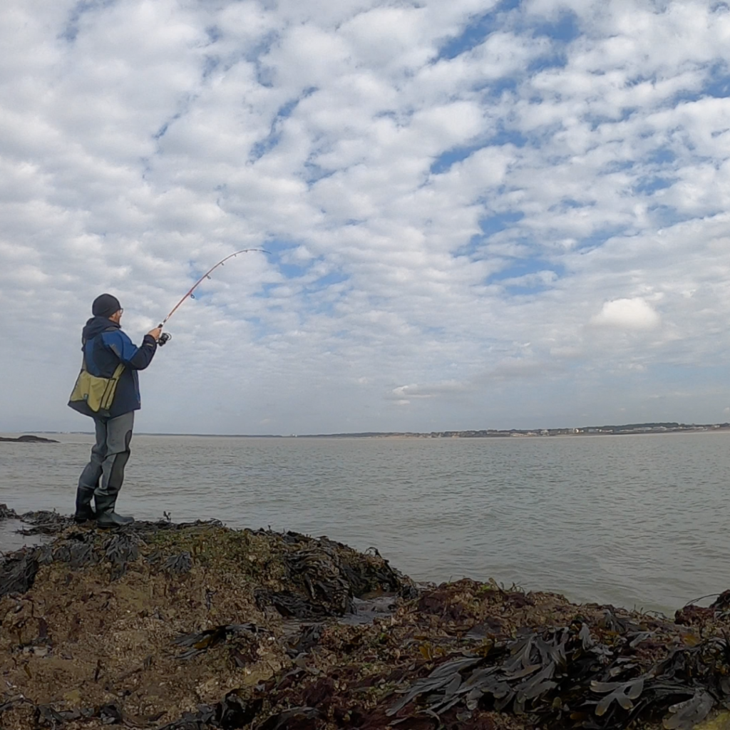 Monsieur Dante YVON, votre moniteur - guide de pêche visible de profil. Celui-ci se tient debout sur des rocher, une canne à pêche à la main. En arrière plan la mer se confond avec le ciel parsemé de nuages. Photo prise lors de la pêche du bar du bord au leurre de surface.