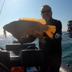 Dante vient de pêcher sur son zodiac un poisson orange avec des points blanc, il est très joli. Il tient le poisson dans ses mains au centre de la photo. Il est sur son zodiac en mer aux alentours de Saint-Michel-Chef-Chef.