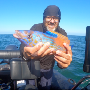 Dante est au centre de la photo, il est sur son zodiac en pleine mer aux alentours de Saint-Michel-Chef-Chef. Il est debout et tient un petit poisson parsemé de bleu vif et de orange, dans ses deux mains, il sourit.