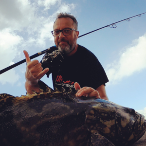 Photo prise en contre plongée. Un poisson se trouve au premier plan, car celui-ci est posé au sol. Dante YVON se tient à l'arrière faisant un signe distinctif des surfeur pour pour exprimer sa joie. Une canne à pêche est également brandit derrière le pêcheur.