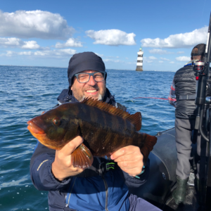 Dante YVON est sur son zodiac en mer aux alentours de Saint-Michel-Chef-Chef. Il est assis et il tient un poisson dans ses deux mains, il est accompagné d'autres pêcheurs que l'on aperçoit sur la gauche.